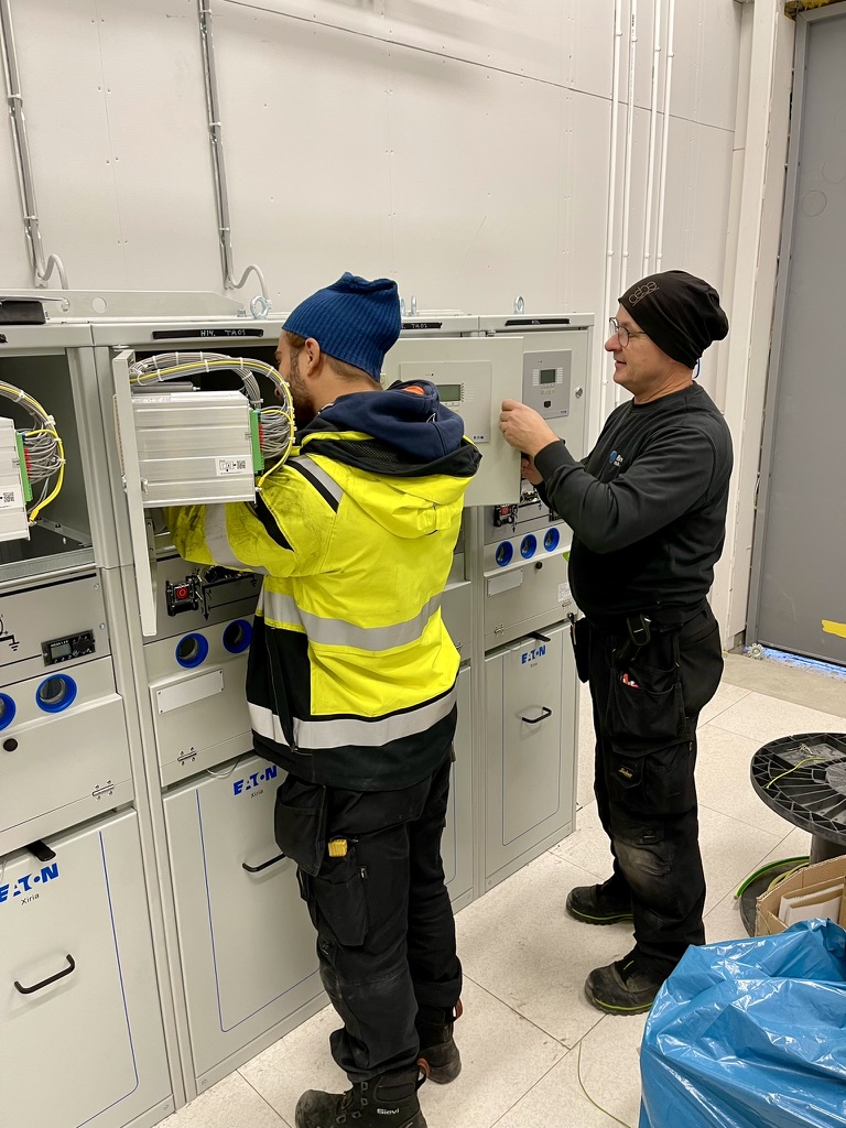 Två montörer arbetar sida vid sida med utrustning i ett ställverk.